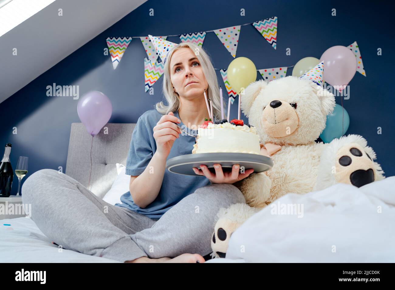 Triste, bouleversée femme en pajama et tête de fête mangeant en cuillère son gâteau d'anniversaire assis sur le lit avec un immense ours en peluche dans la chambre décorée. Fête le bi Banque D'Images Triste, bouleversée femme en pajama et tête de fête mangeant en cuillère son gâteau d'anniversaire assis sur le lit avec un immense ours en peluche dans la chambre décorée. Fête le bi Banque D'Images