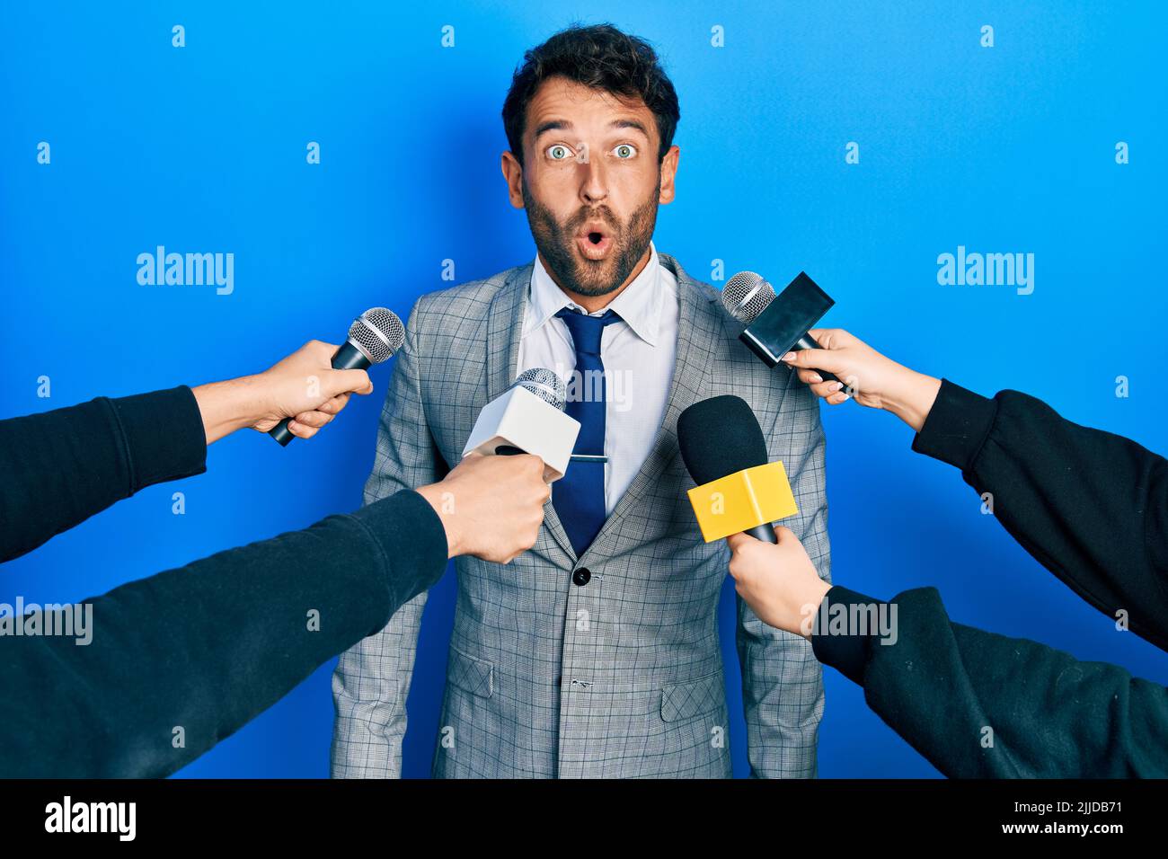 Beau homme d'affaires avec la barbe étant interviewé par des reporters tenant le microphone effrayés et surpris avec la bouche ouverte pour la surprise, le visage incrédulé Banque D'Images
