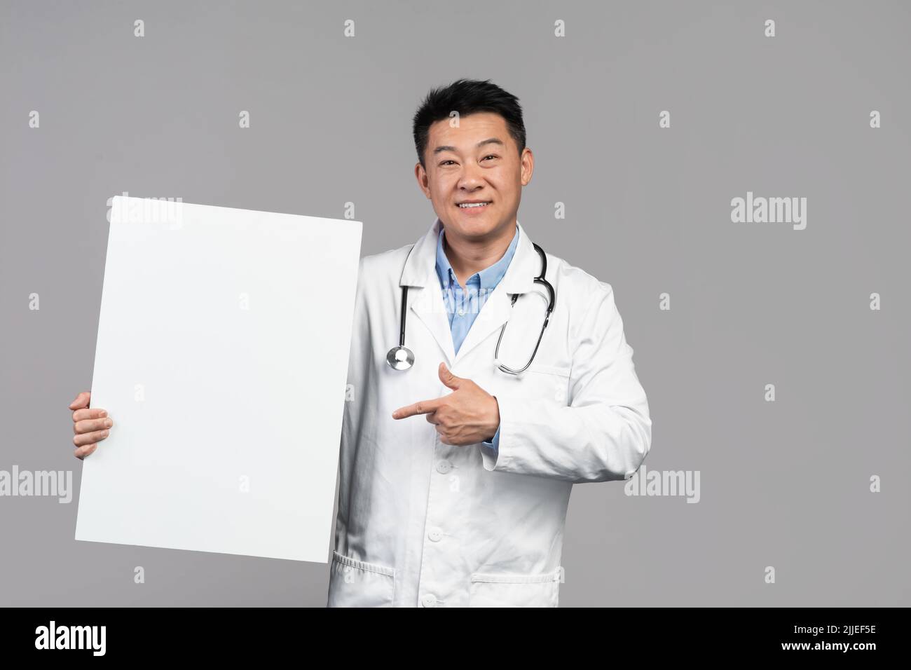 Souriant beau adulte asiatique homme médecin en manteau blanc avec stéthoscope pointe le doigt sur la bannière Banque D'Images