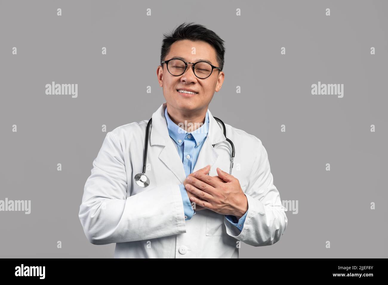 Joyeux millennium coréen gars docteur en manteau blanc, lunettes avec stéthoscope appuie les mains au coeur Banque D'Images