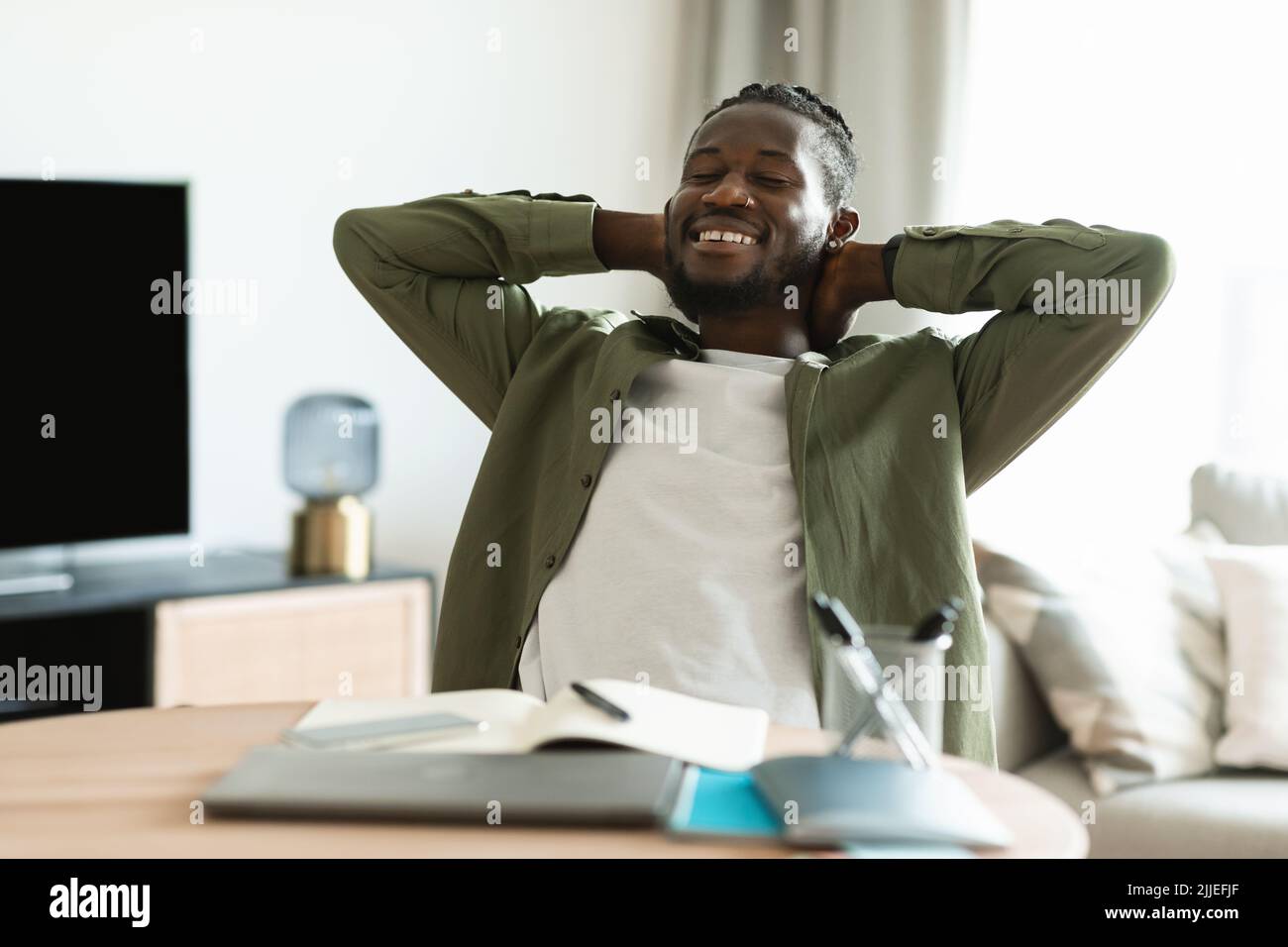 Heureux afro-américain avec les yeux fermés assis à la table et penché en arrière dans la chaise, appréciant le travail indépendant Banque D'Images