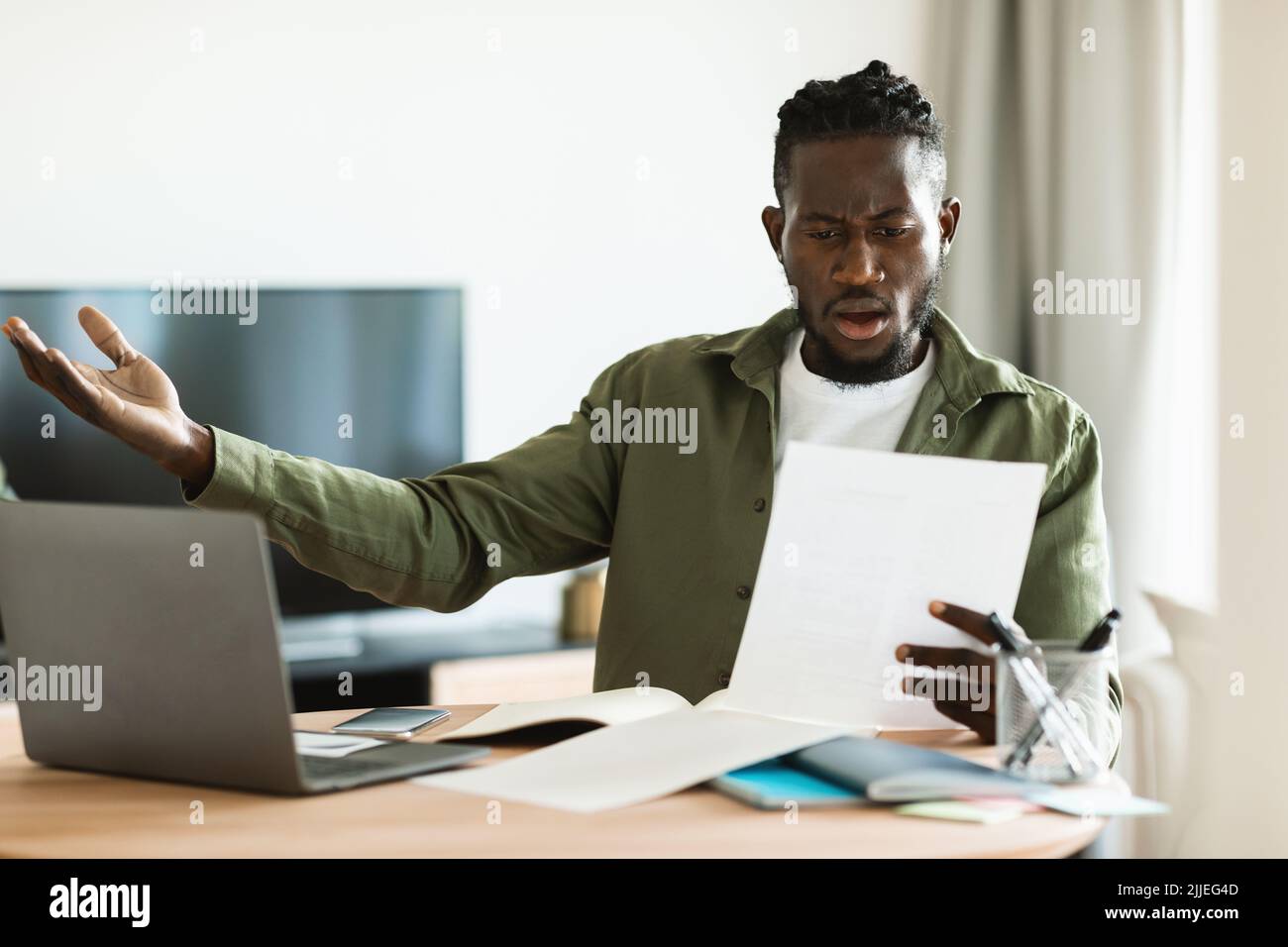 Homme noir choqué lisant le courrier papier et se propageant les mains, assis à la table avec un ordinateur portable au bureau à domicile Banque D'Images
