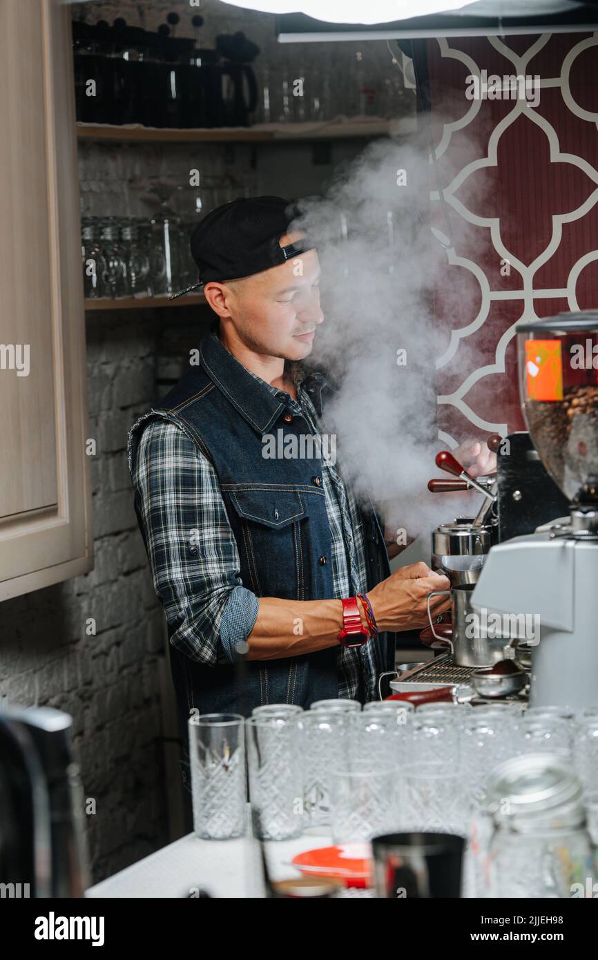 Un barista masculin prépare du café dans une cuisine à café vapeur. Il porte une chemise à carreaux, une veste sans manches et un chapeau de soleil à l'envers. Banque D'Images