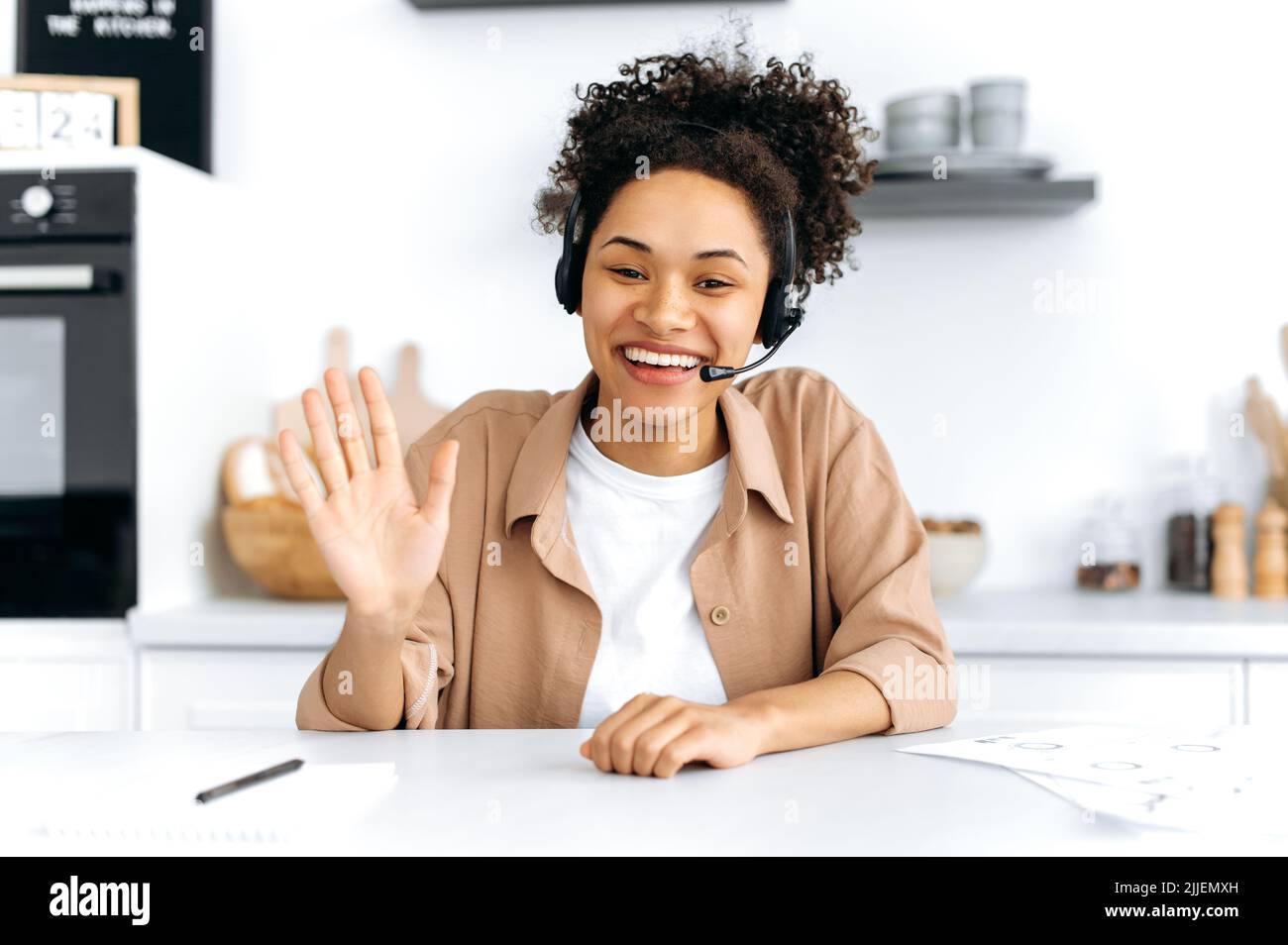 Positive confiante jeune femme afro-américaine aux cheveux bouclés avec micro-casque, employé de centre d'appel, consultant, parle par vidéoconférence, femme mentor enregistrement webinaire, conférence, gestuelle avec les mains, sourire Banque D'Images