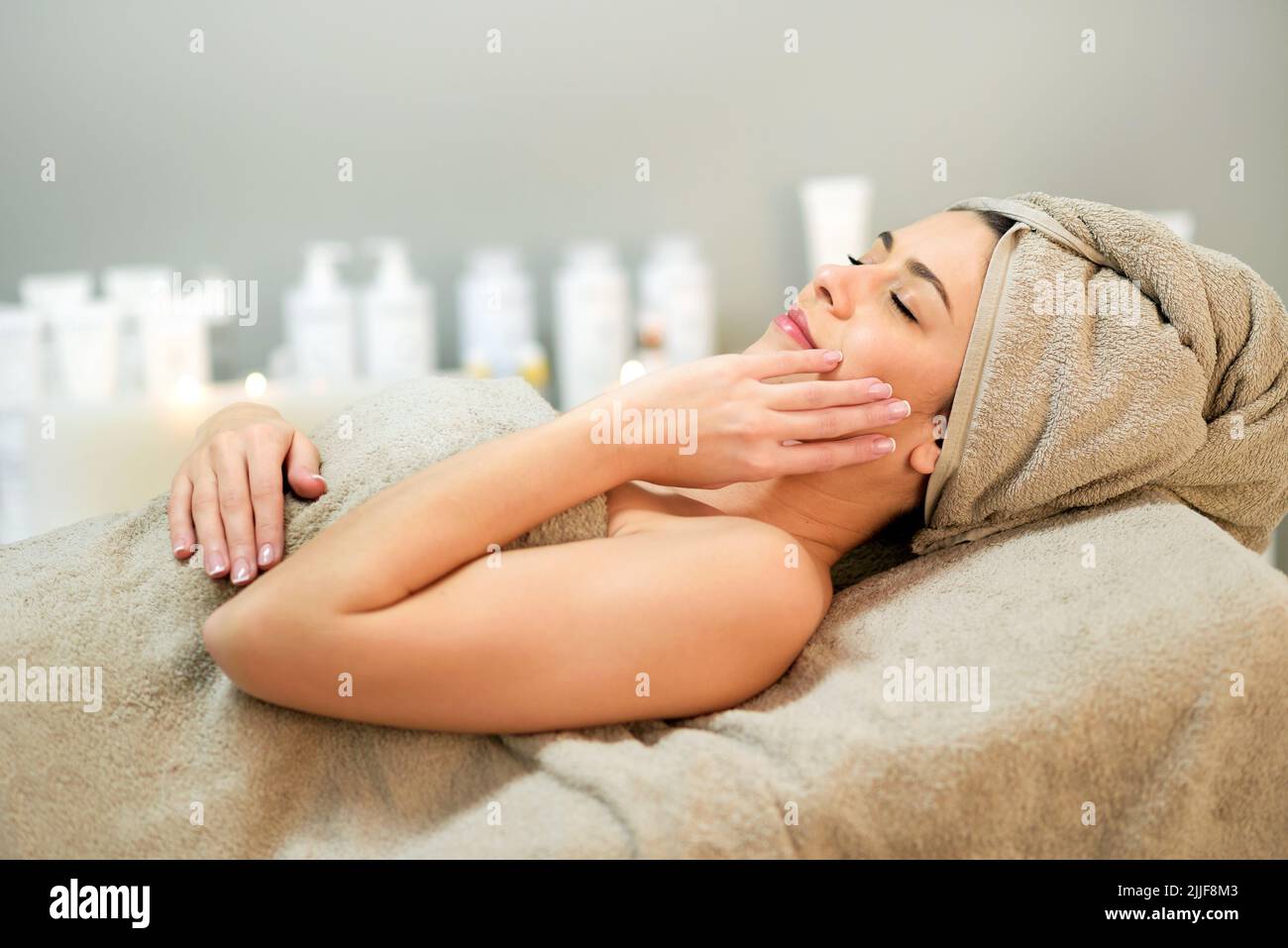 Vue latérale d'une femme détendue enveloppée d'une serviette touchant la joue en étant allongé sur le canapé pendant la procédure de spa dans le salon Banque D'Images