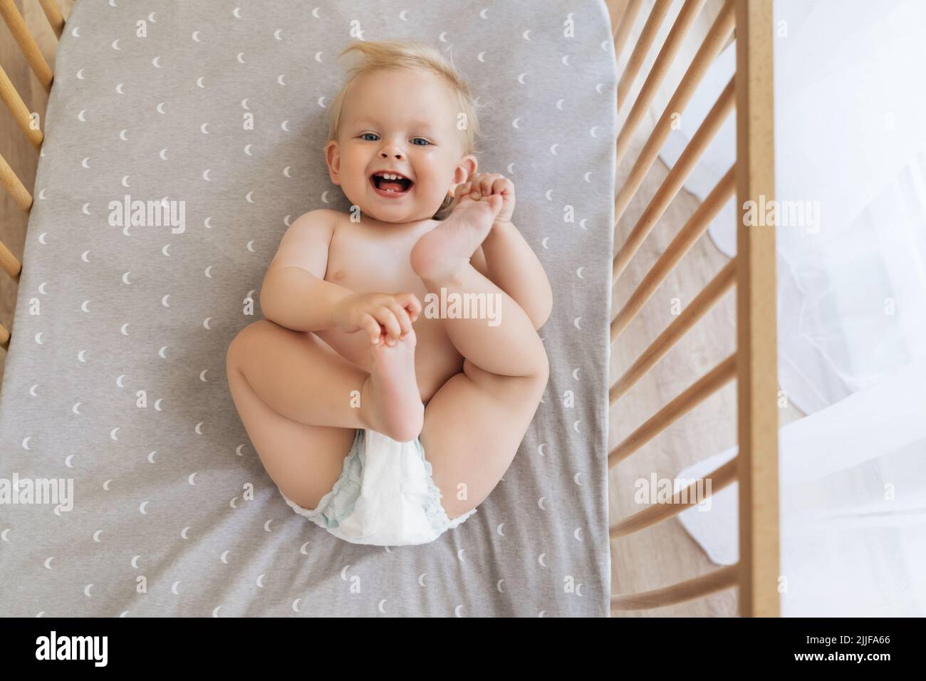 Portrait en haut de l'image de bébé amusant et amusant mignon en couche allongé sur son dos en jouant avec les pieds, en riant avec l'appareil photo dans le lit sur un drap gris avec un imprimé demi-lune Banque D'Images