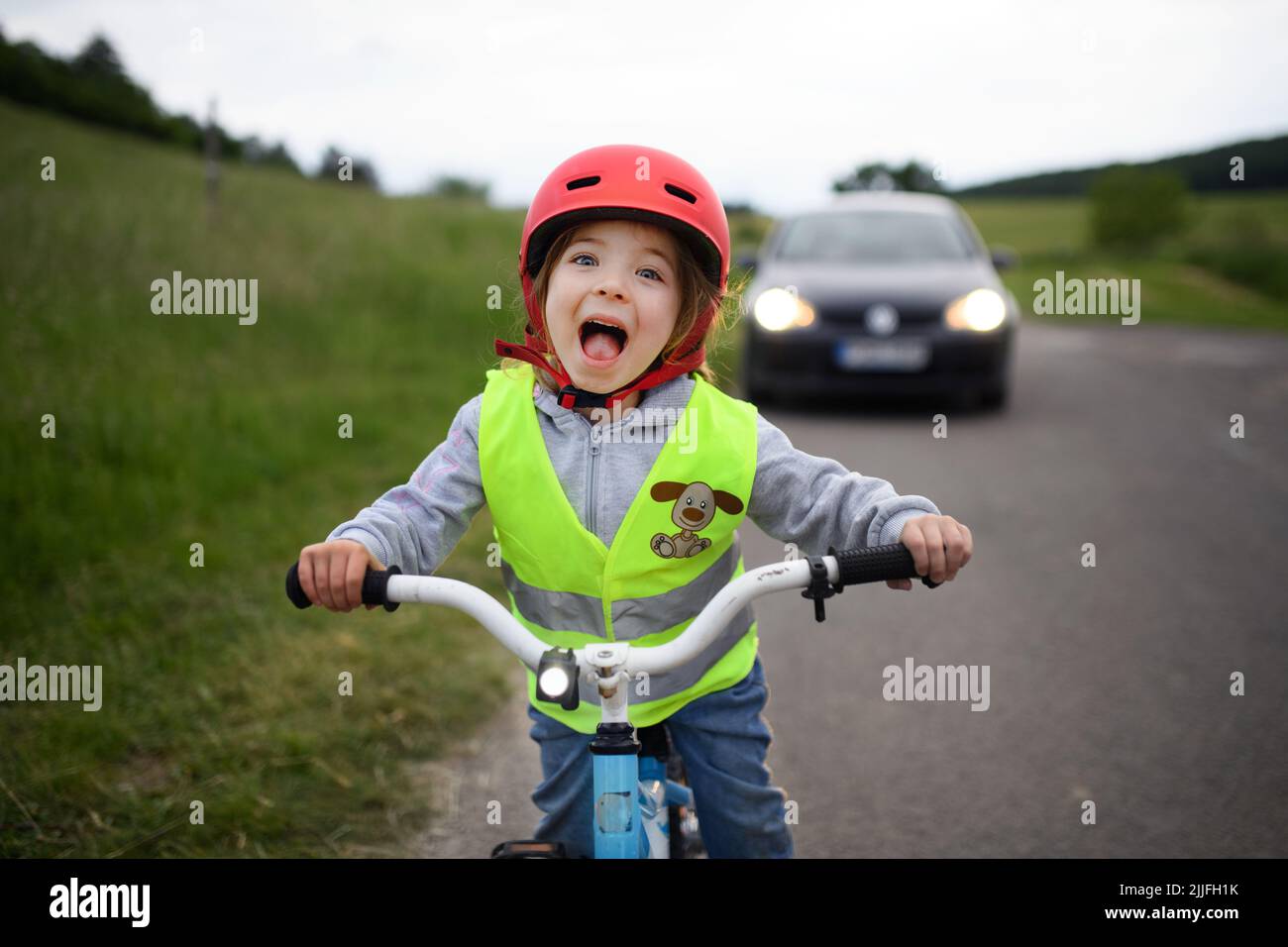 Portrait d'une petite fille excitée dans un gilet réfléchissant à vélo sur la route avec une voiture derrière elle, concept d'éducation à la sécurité routière. Banque D'Images