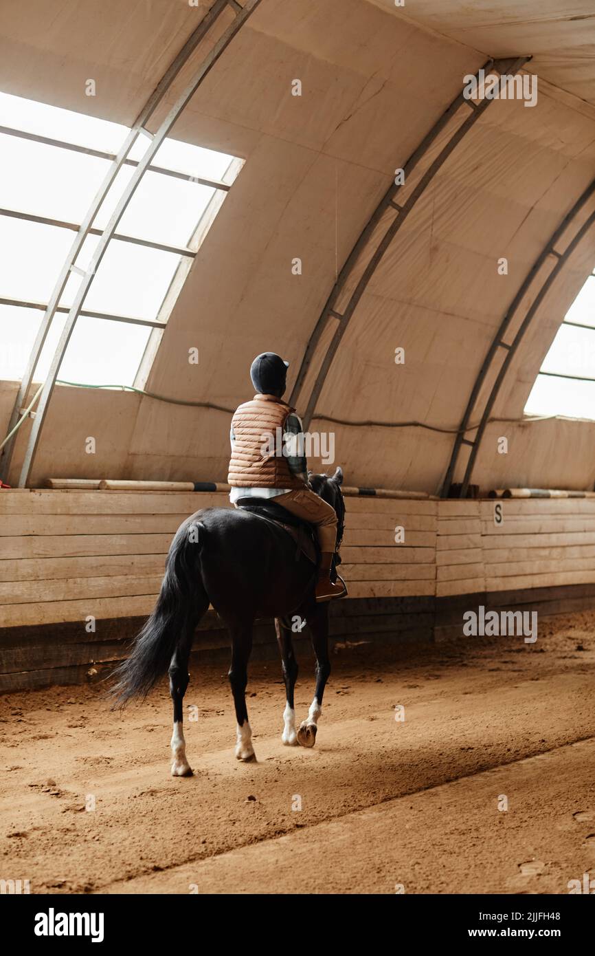 Vue arrière verticale sur une jeune femme à cheval dans une arène intérieure dans un ranch ou un stade d'entraînement Banque D'Images