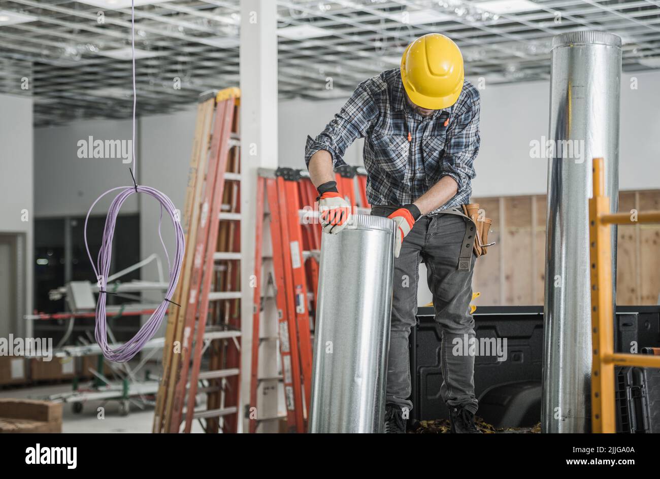 Entrepreneur caucasien en chapeau dur jaune déplaçant le tuyau de ventilation pendant l'installation du système de contrôle de température HVAC dans l'entrepôt commercial Banque D'Images