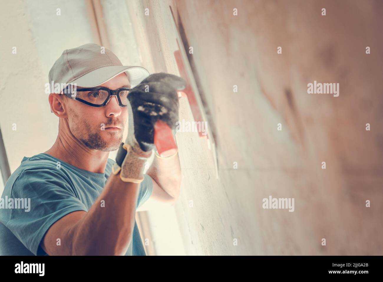 Gros plan d'un travailleur caucasien dans des lunettes de sécurité remplissage et lissage soigneux du mur avec de la putty de Gypsum pendant les travaux de rénovation dans la maison résidentielle. Banque D'Images
