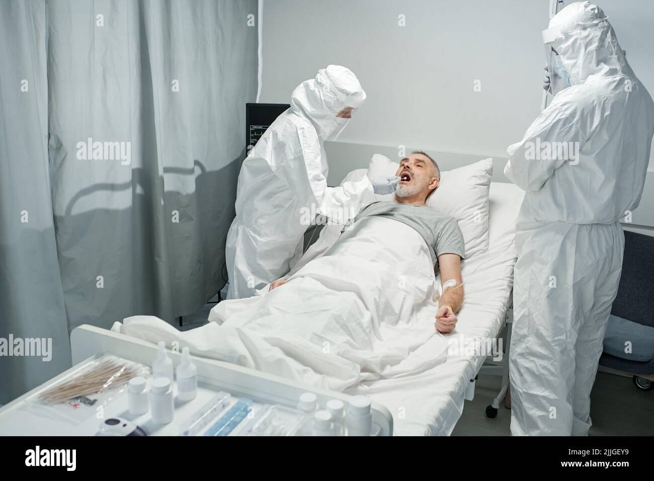 Médecins méconnaissables portant des combinaisons de protection blanches avec des masques qui testent l'homme mature pour des maladies infectieuses ou bactériennes Banque D'Images