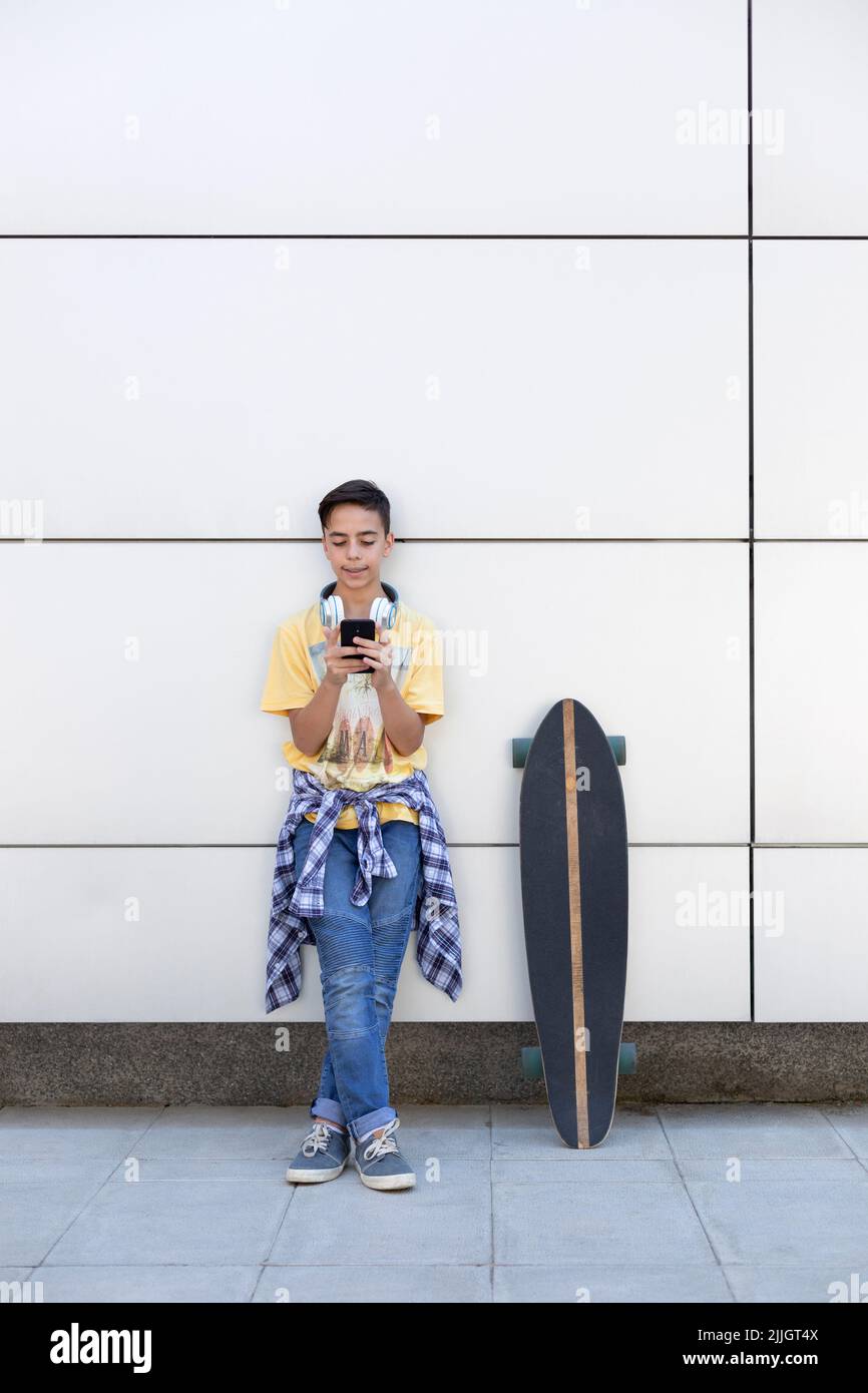 Adolescent de race blanche avec skateboard à l'aide d'un téléphone portable. Il est isolé sur un mur. Espace pour le texte. Banque D'Images