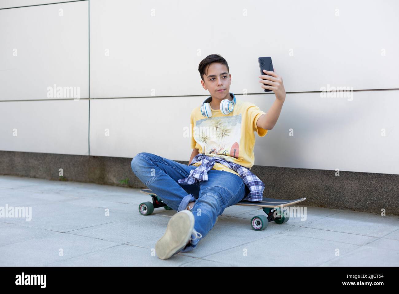 Adolescent caucasien assis sur un skateboard prenant une photo de lui-même avec un smartphone. Style de vie urbain. Espace pour le texte. Banque D'Images