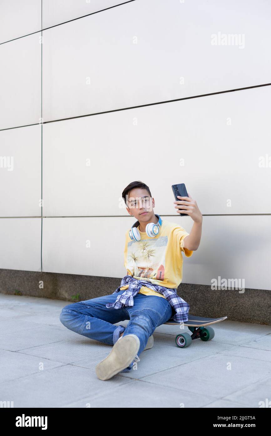 Adolescent caucasien assis sur un skateboard prenant une photo de lui-même avec un téléphone portable. Style de vie urbain. Espace pour le texte. Banque D'Images