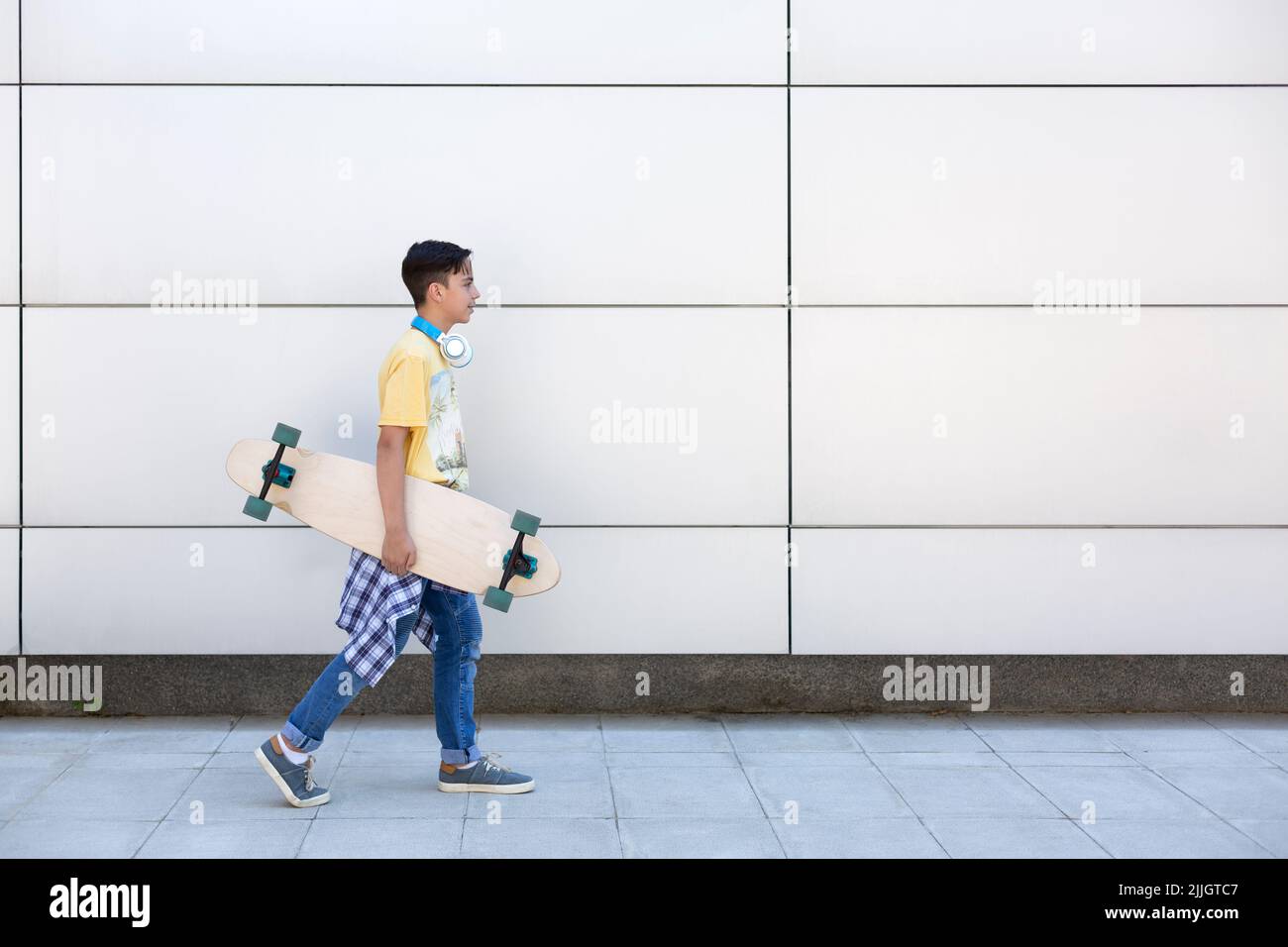 Joyeux adolescent caucasien avec un long skateboard dans sa main marchant à côté d'un mur. Style de vie urbain. Espace pour le texte. Banque D'Images
