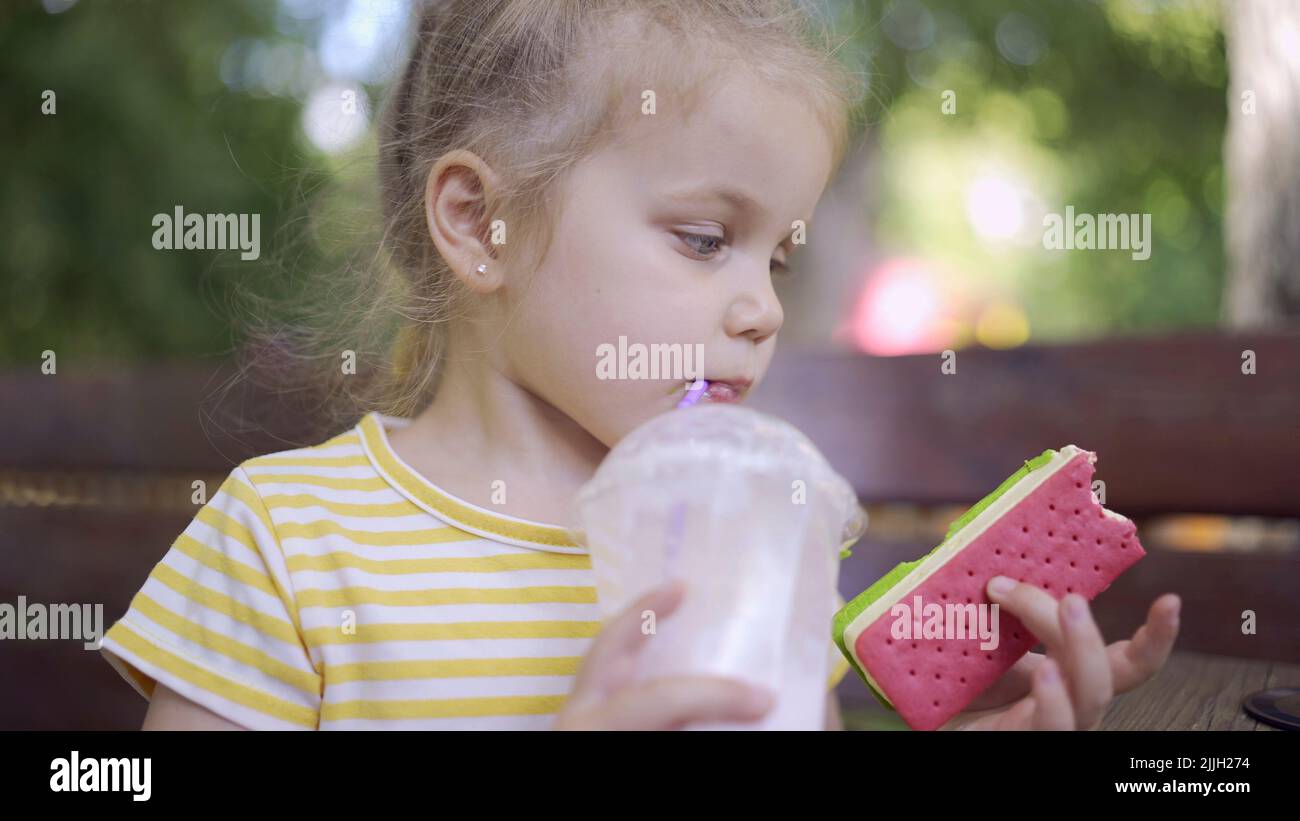 Une petite fille boit un milkshake à travers une paille et saumone à la glace de son autre main. Portrait en gros plan d'une petite fille adorable assise sur le banc de parc a Banque D'Images