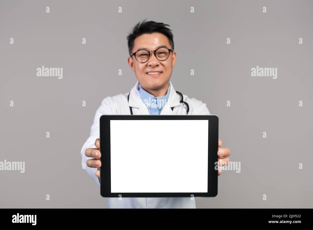 Joyeux médecin japonais adulte confiant en manteau blanc et des lunettes montre le comprimé avec écran blanc Banque D'Images