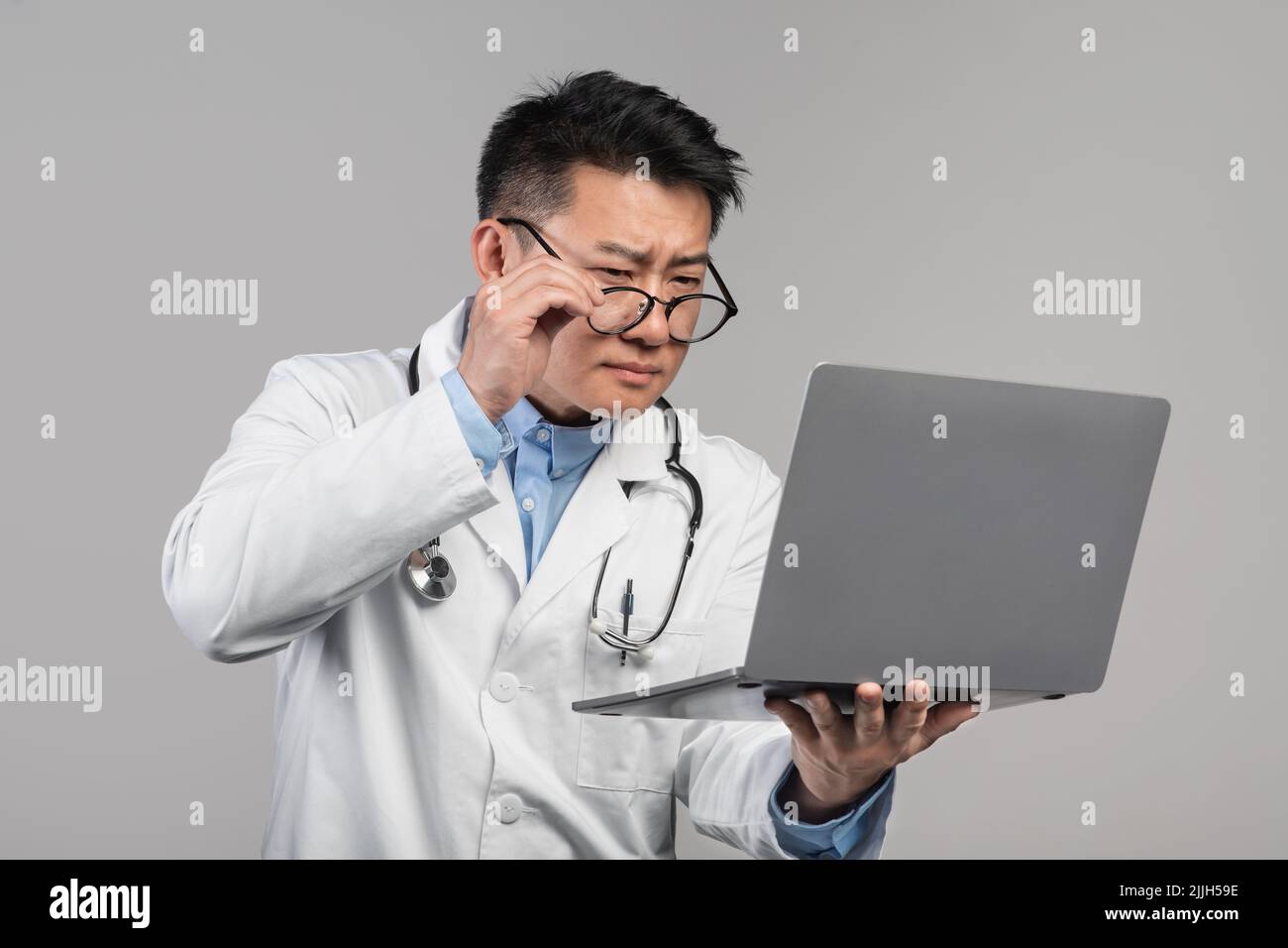 Confus adulte concentré asiatique homme médecin dans le manteau blanc prend des lunettes, regarde l'ordinateur portable Banque D'Images