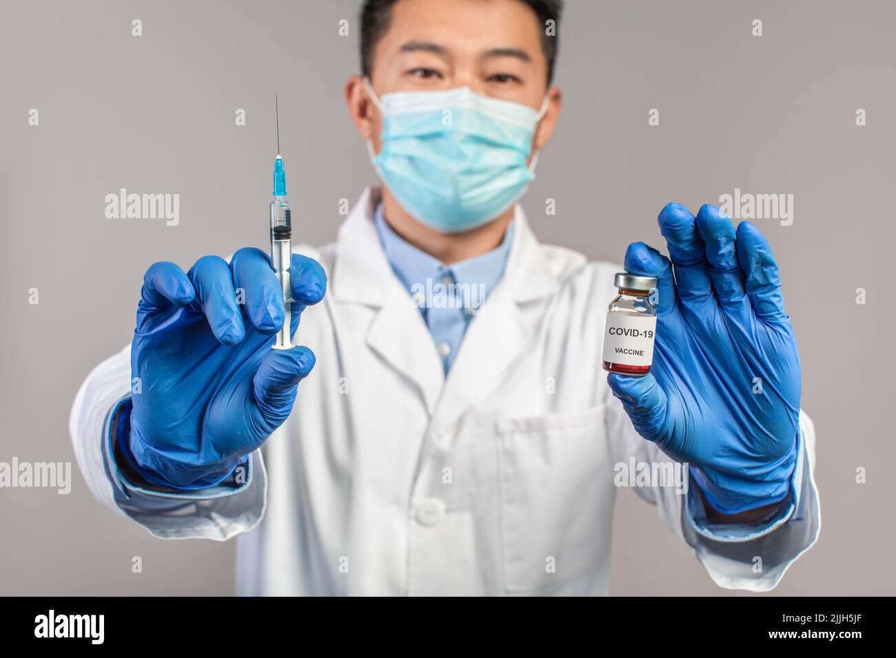 Un médecin chinois sérieux adulte en manteau blanc, masque de protection, gants montre une seringue et un vaccin Banque D'Images