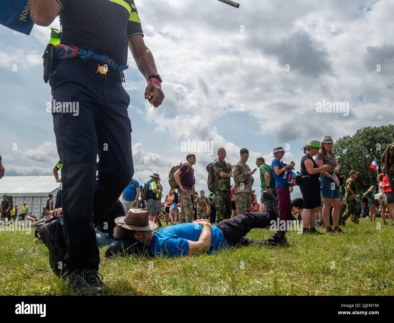 Un homme repose sur l'herbe tandis que d'autres participants marchent à côté de lui pendant le plus grand événement de marche de plusieurs jours au monde. La marche des quatre jours (en néerlandais 'Vierdaagse') est considérée comme le premier exemple de l'esprit sportif et des liens internationaux entre militaires et civils de nombreux pays différents. Après deux ans d'annulation, il a été retenu à nouveau, mais le premier jour a été annulé en raison de températures chaudes, transformant les quatre marches en trois jours de marche. Il s'agit de l'édition 104 et le total officiel des marcheurs enregistrés était de 38 455 dans 69 pays. Ils peuvent choisir de marcher 30km, 40km, ou Banque D'Images