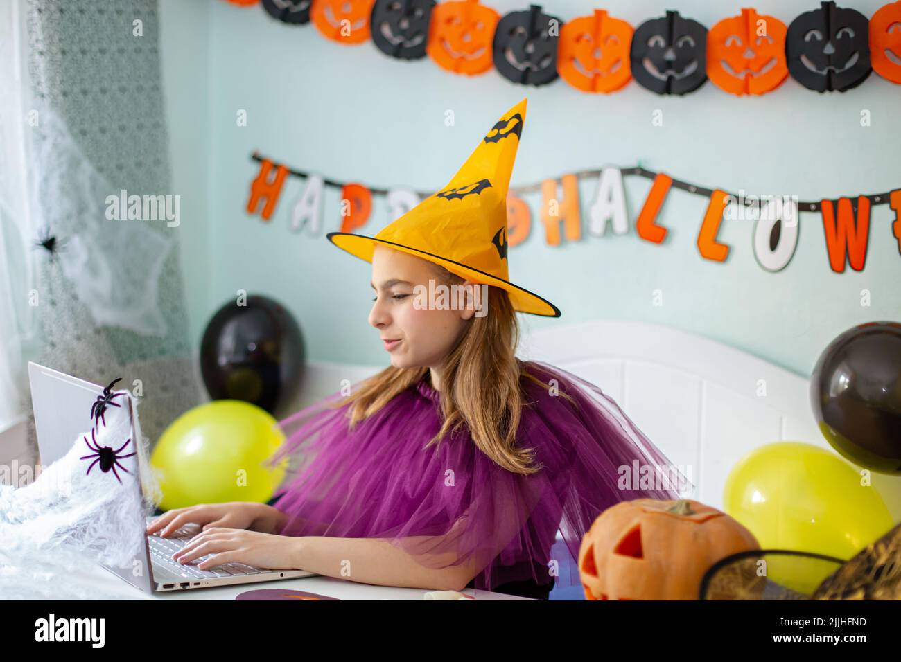 Jolie petite fille portant un chapeau de sorcière assis derrière une table dans la chambre décorée sur le thème d'Halloween. Concept de fête d'Halloween. Préparation pour la célébration, Banque D'Images