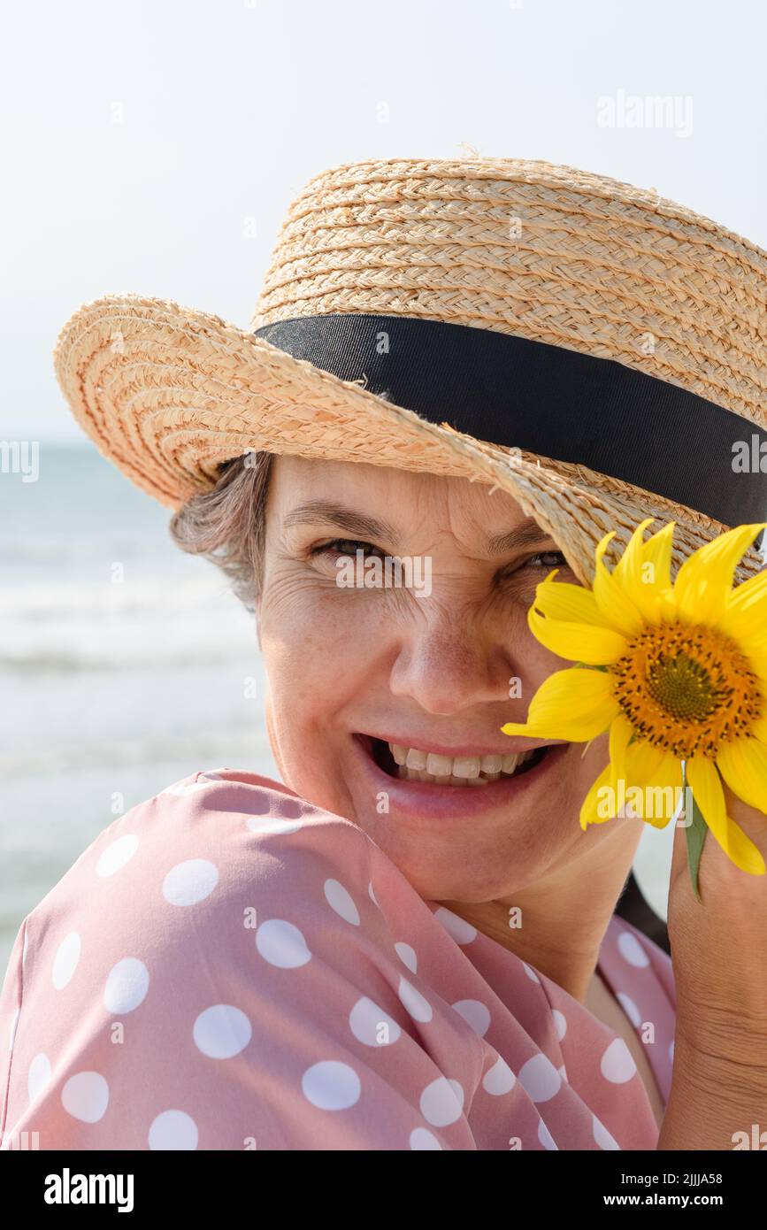 Femme aux cheveux gris souriants dans un boater et une robe rose avec des pois blancs tenant une fleur jaune regardant l'appareil photo, gros plan portrait. Banque D'Images