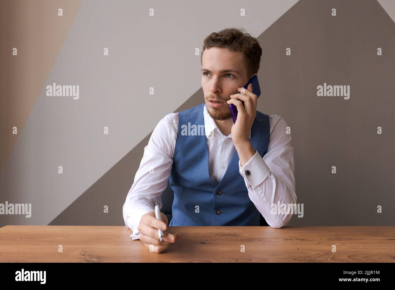 Un jeune homme d'affaires sérieux à barbe en chemise blanche et gilet bleu est assis à table au bureau au travail et parle sur son téléphone portable. Le responsable des travailleurs caucasiens travaille avec les clients, les appels à froid Banque D'Images