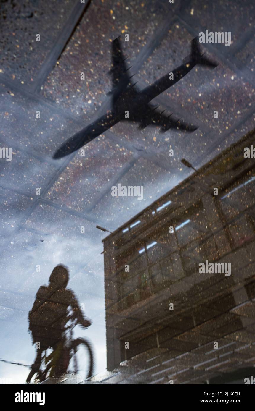 Silhouette de réflexion floue d'une personne sur une promenade à vélo seule sur un trottoir humide de la ville le jour des pluies. L'avion vole à travers le ciel. Photo abstraite Banque D'Images