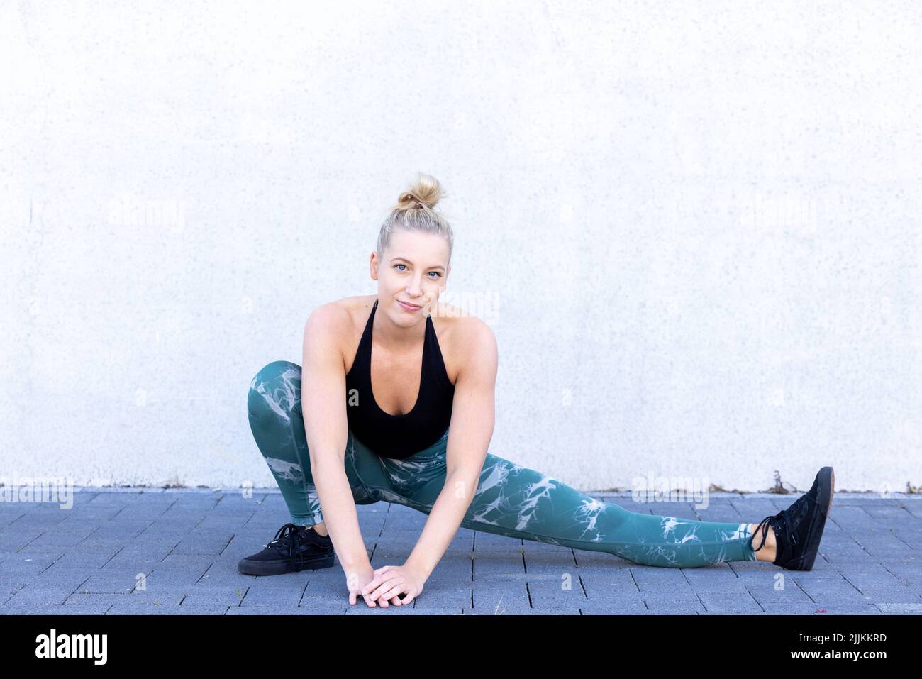 Belle jeune femme blonde de forme physique portant des vêtements de sport noirs et bleus s'étirant et réchauffant quadriceps et fessiers, fesses, sur les jambes le matin. Femme sportive effectuant un entraînement d'échauffement par un mur. Photo de haute qualité Banque D'Images