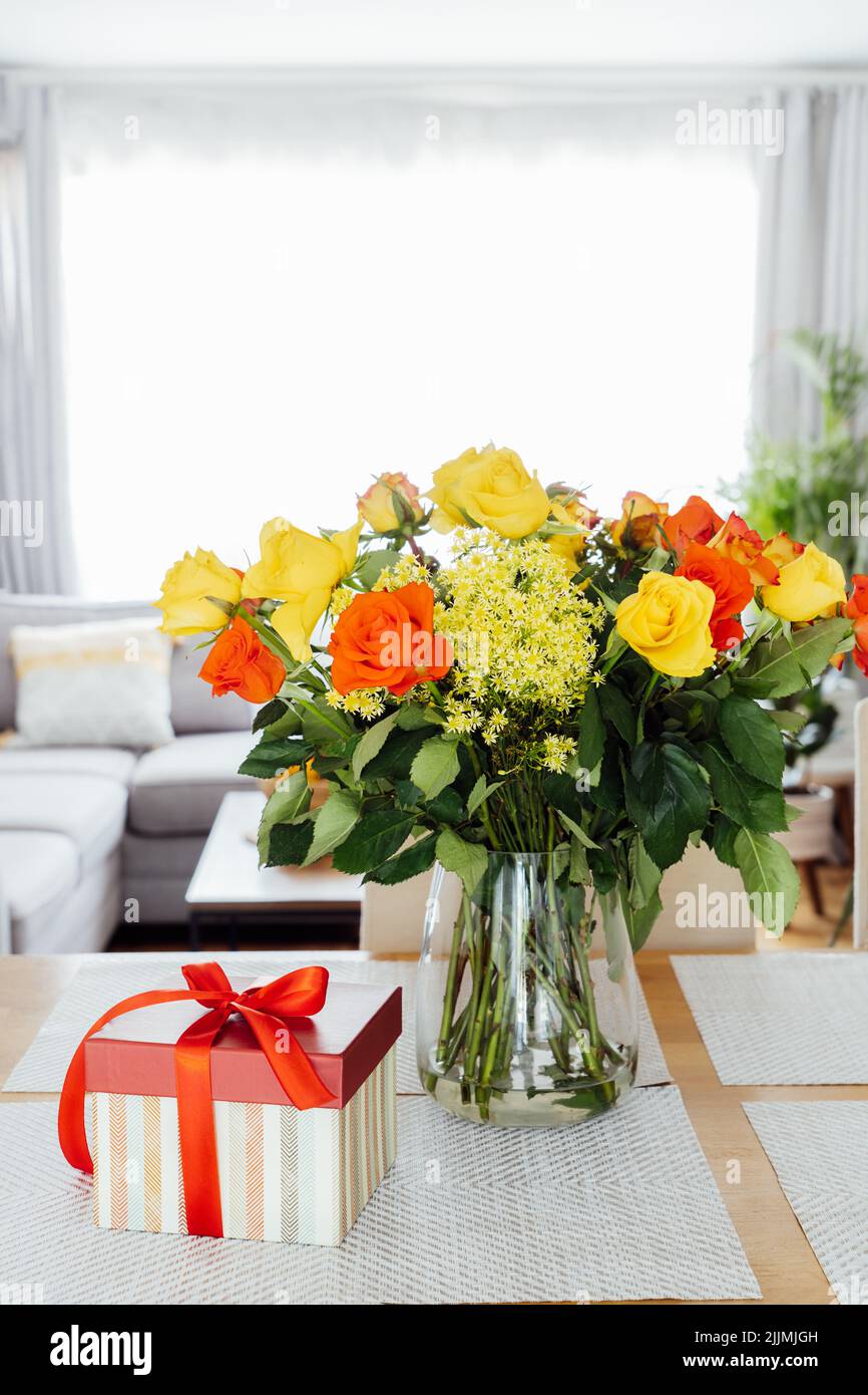 Roses jaunes et oranges dans un vase et boîte cadeau avec ruban rouge sur la table de comptoir de cuisine avec espace ouvert salon arrière-plan. Cadeau pour Thanksgiving Banque D'Images Roses jaunes et oranges dans un vase et boîte cadeau avec ruban rouge sur la table de comptoir de cuisine avec espace ouvert salon arrière-plan. Cadeau pour Thanksgiving Banque D'Images