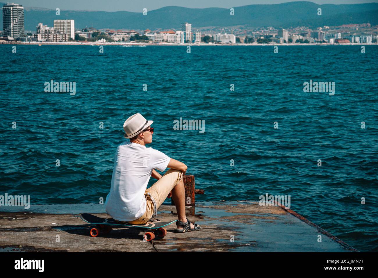 Un jeune caucasien en lunettes de soleil assis sur un skateboard sur un quai en béton qui regarde une mer Banque D'Images