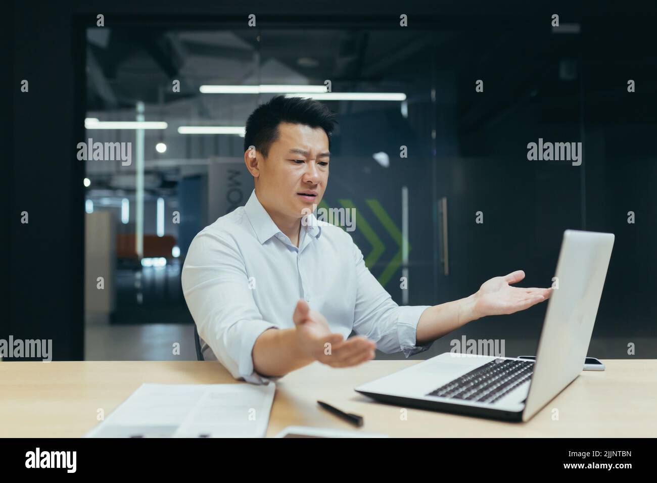 Un jeune homme beau, un homme d'affaires asiatique confus, assis à un ordinateur portable dans le bureau, parle sur un appel vidéo, répand ses mains, montre qu'il ne comprend pas. Dans un bureau moderne. Banque D'Images