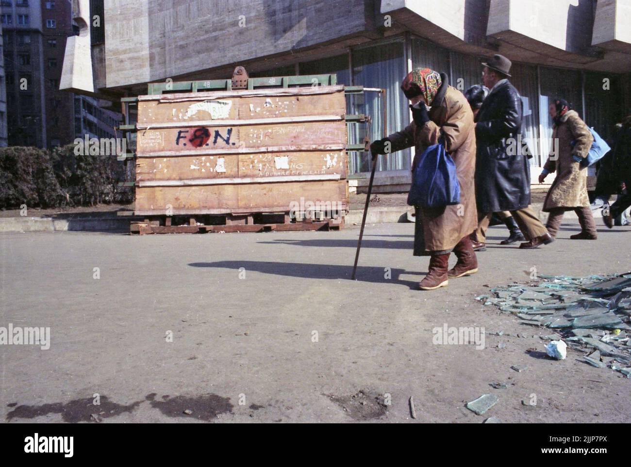 Bucarest, Roumanie, janvier 1989. Les gens passent par des brisures de verre et de débris sur la place de l'université, à côté de l'hôtel Intercontinental, quelques jours après la révolution anticommuniste de décembre 1989. Une femme âgée couvre son visage avec un mouchoir. Sur le panneau en bois quelqu'un a écrit 'F.S.N.' (Front du salut national - le parti qui a pris le pouvoir après l'élimination de Ceausescu), avec le marteau et la faucille communistes dedans, faisant allusion à l'inclination du parti. Banque D'Images