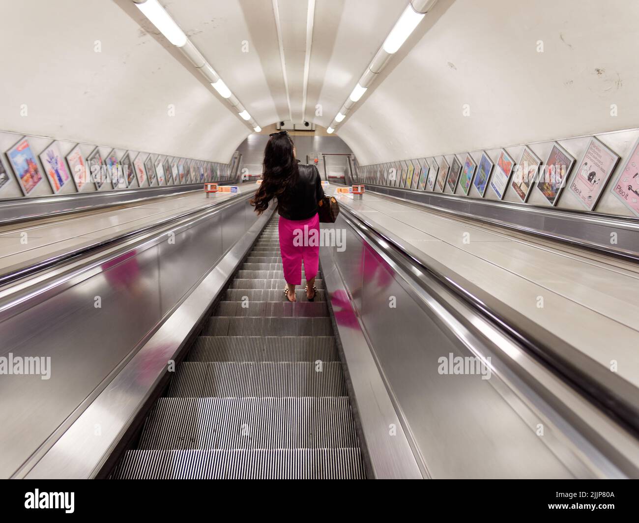 Londres, Grand Londres, Angleterre, 02 juillet 2022 : Dame avec un pantalon rose et une veste noire sur un escalier roulant sur une station de métro autrement vide Banque D'Images