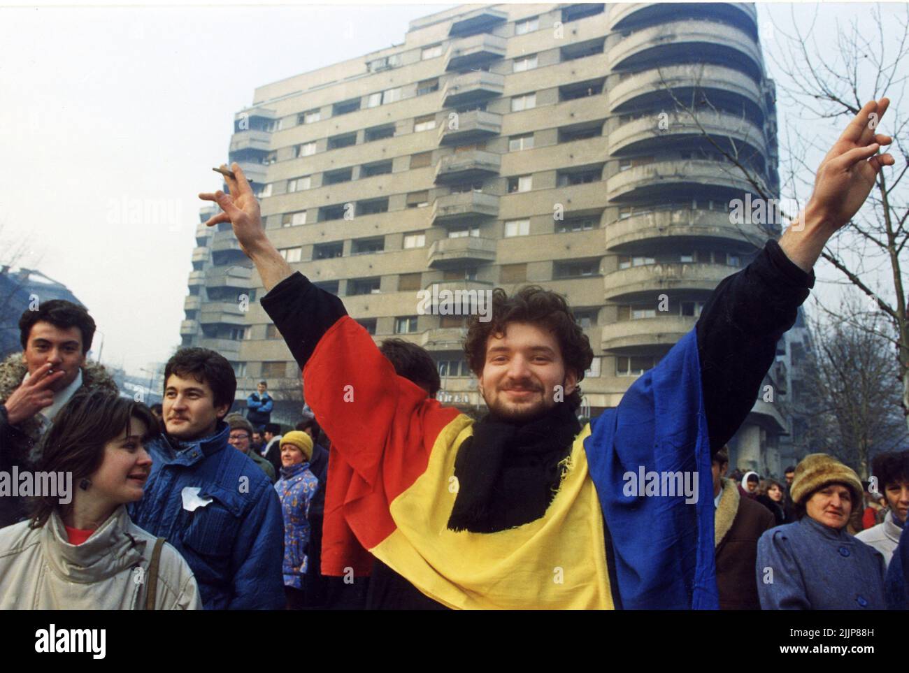 Bucarest, Roumanie, janvier 1990. Après la révolution anticommuniste de décembre 1989, les gens se rassembleraient tous les jours sur la place de l'université pour protester contre les nouveaux peuples au pouvoir, qui étaient les anciens responsables communistes. Un jeune homme fait le signe de la victoire, portant le drapeau national avec l'emblème socialiste coupé, un symbole pendant la révolution, représentant la fin de l'ère communiste. Banque D'Images