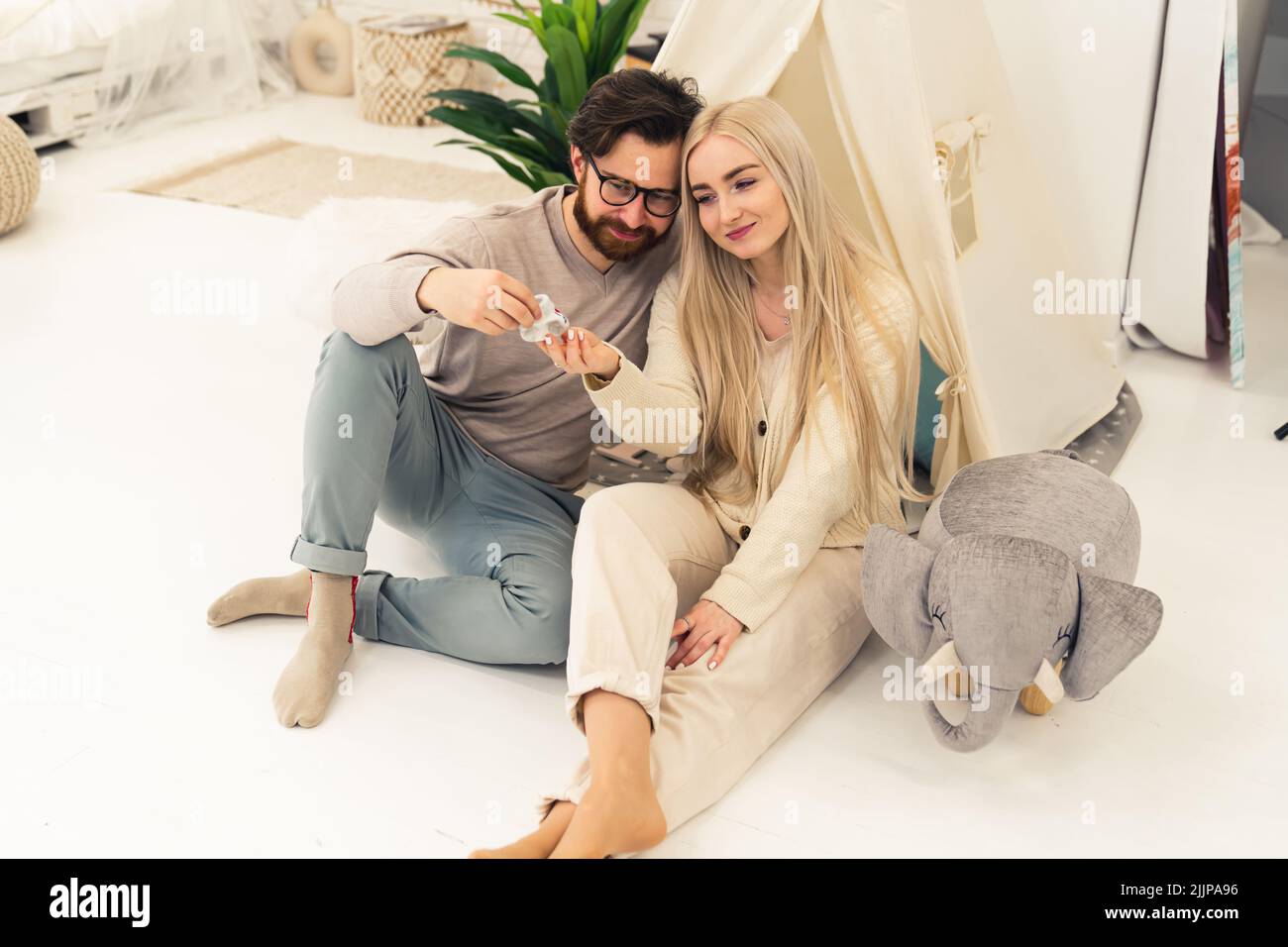 Jeune s'attendant à un couple blanc regardant affectueusement la chaussure de bébé tricotée sur le sol à côté de la tente tipi et de la pluschie d'éléphant. Prise de vue en intérieur. Photo de haute qualité Banque D'Images