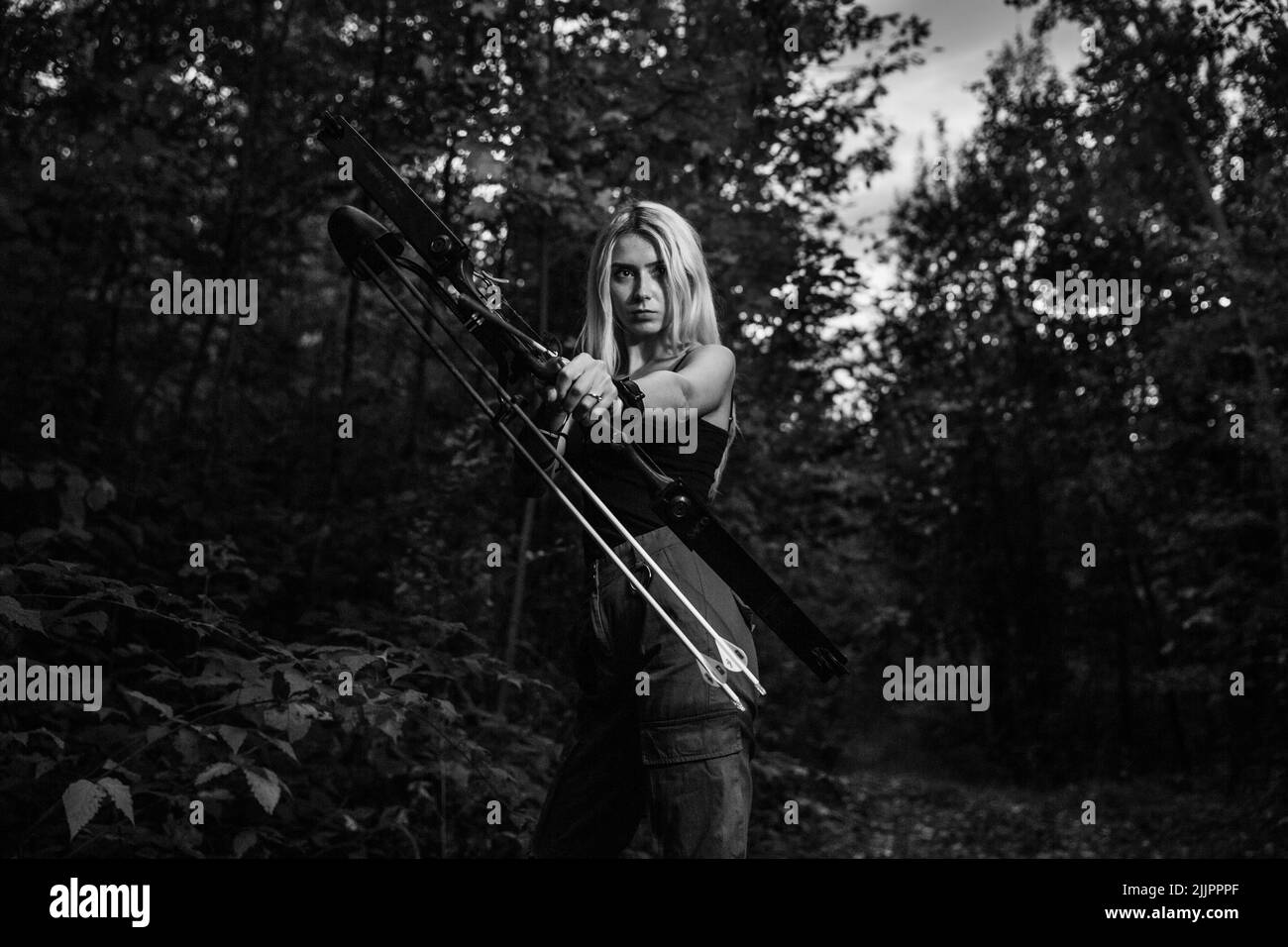 Portrait en niveaux de gris d'une femme blonde caucasienne tenant une flèche et une lance dans une forêt Banque D'Images