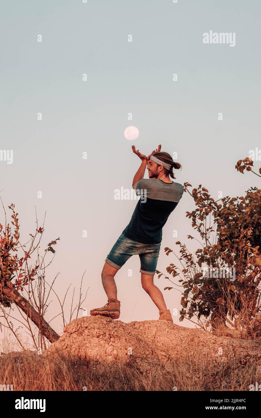 Photo verticale d'un homme caucasien debout sur un rocher essayant de prendre la lune avec ses mains sur un fond ciel clair Banque D'Images