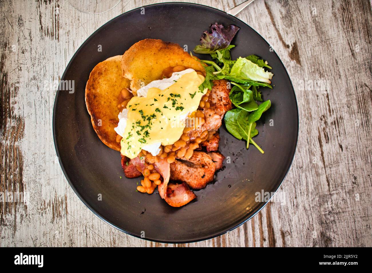 Vue de dessus de l'œuf Benedict servi avec du pain frit, du bacon et de la laitue sur une assiette noire Banque D'Images Vue de dessus de l'œuf Benedict servi avec du pain frit, du bacon et de la laitue sur une assiette noire Banque D'Images