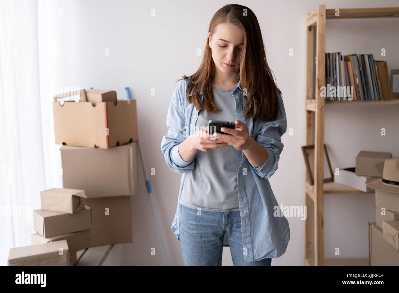 une jeune femme sérieuse se déplace, se tient dans des boîtes en carton, utilise un smartphone, se déplace dans une nouvelle maison, se déplace jour et la livraison d'appel, en ligne Banque D'Images