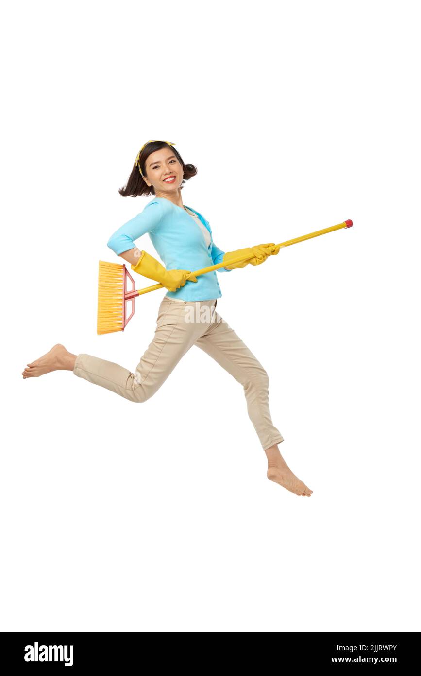 Femme asiatique énergique portant des gants en caoutchouc sautant dans l'air avec un balai et regardant l'appareil photo avec un sourire crasseux tout en étant distrait du sol, isolé sur fond blanc Banque D'Images
