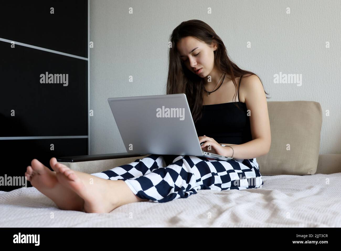 Jolie jeune fille avec de longs cheveux portant noir dessus assis avec un ordinateur portable sur le canapé. Concept de travail, d'étude ou de loisirs à la maison Banque D'Images