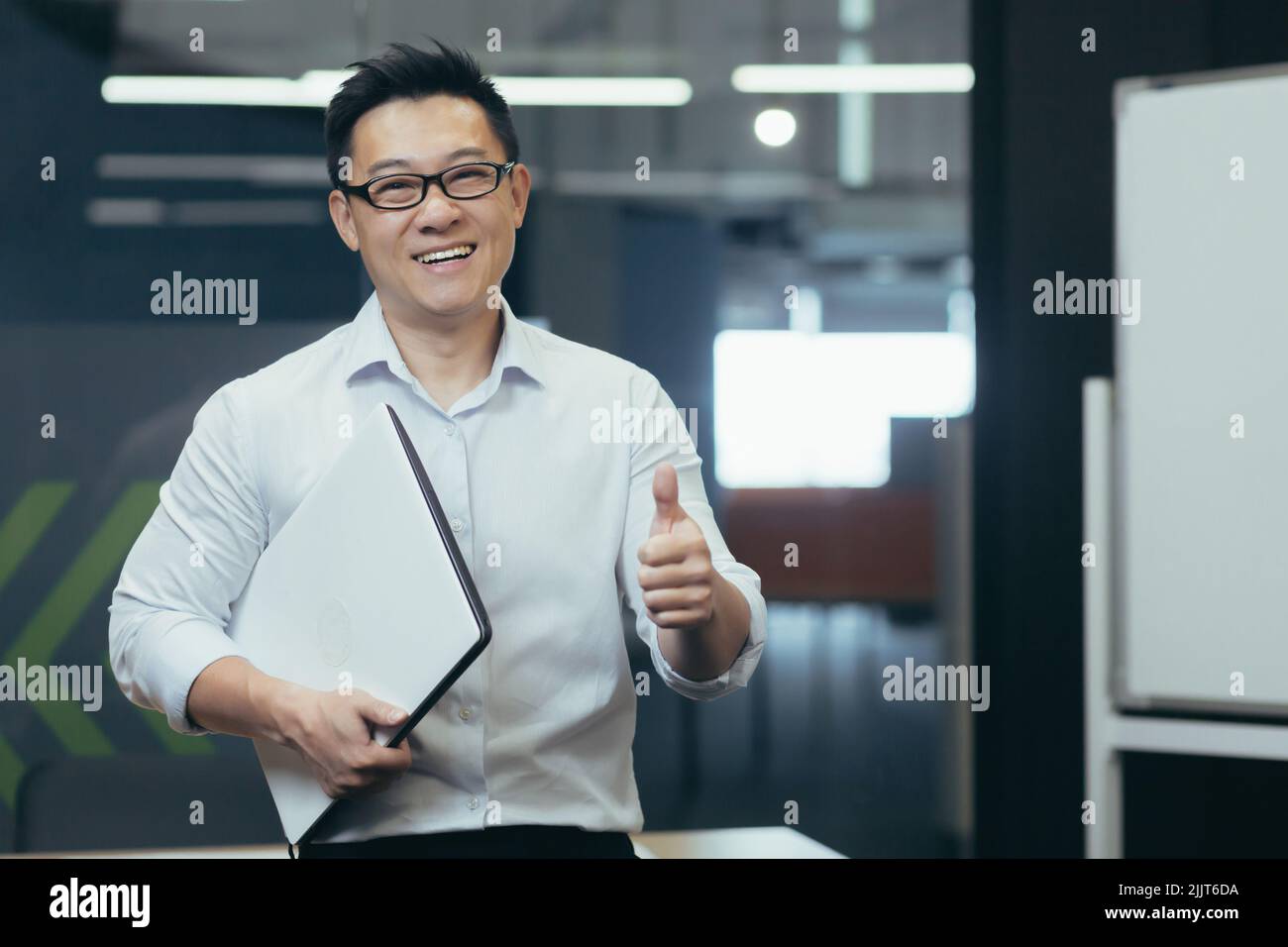 Portrait d'un jeune asiatique stagiaire, assistant au bureau. Il est debout dans des lunettes, pointant ok, tenant un dossier dans ses mains, souriant, regardant la caméra. Banque D'Images