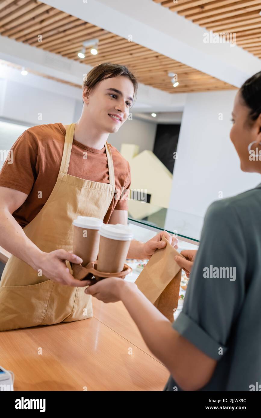 Vendeur souriant donnant du café à emporter et sac en papier à un client afro-américain flou dans la confiserie Banque D'Images