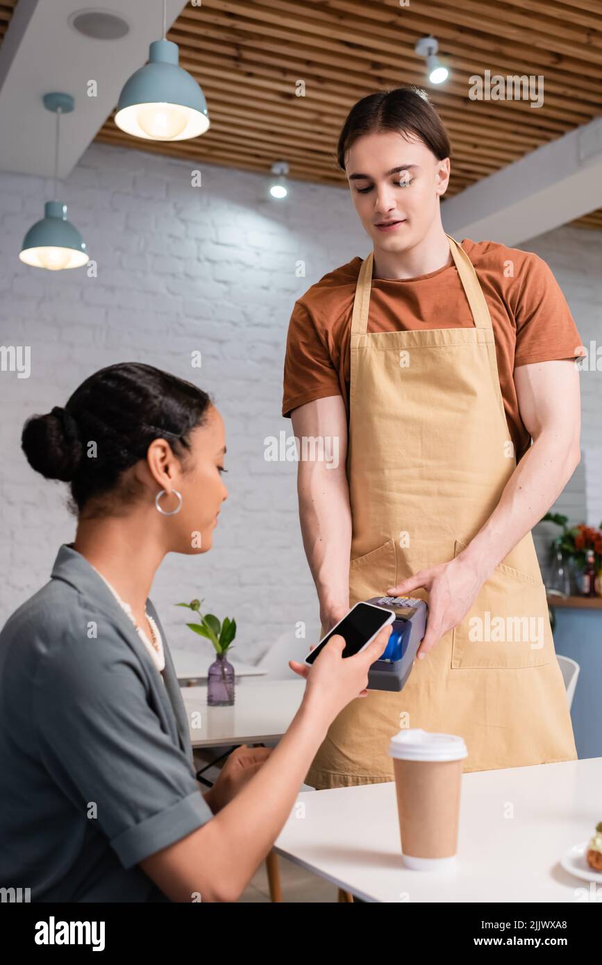 Vendeur détenant un terminal de paiement près d'un client afro-américain avec un smartphone et du café dans des confiseries Banque D'Images