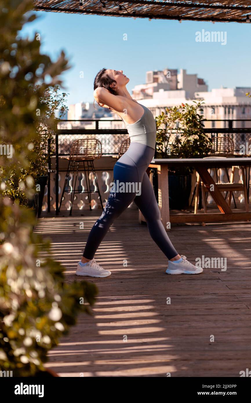 Vue latérale du corps de la femme active dans les vêtements de sport faire  de l'exercice avec la jambe relevée tout en s'exerçant sur la terrasse du  bâtiment pendant l'entraînement le jour