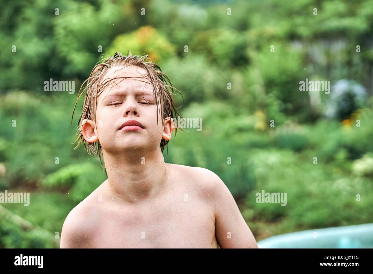 L'écolier mignon aux cheveux blonds humides se dresse contre les arbres luxuriants et la verdure en campagne. Portrait d'un garçon décontracté en gros plan Banque D'Images