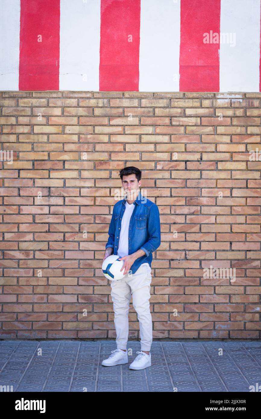 Portrait complet d'un jeune homme beau debout avec un ballon de football tout en se tenant contre un mur de briques Banque D'Images