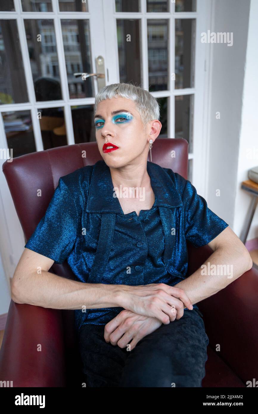 Portrait d'un homme androgyne confiant portant un maquillage assis sur un fauteuil à la maison Banque D'Images