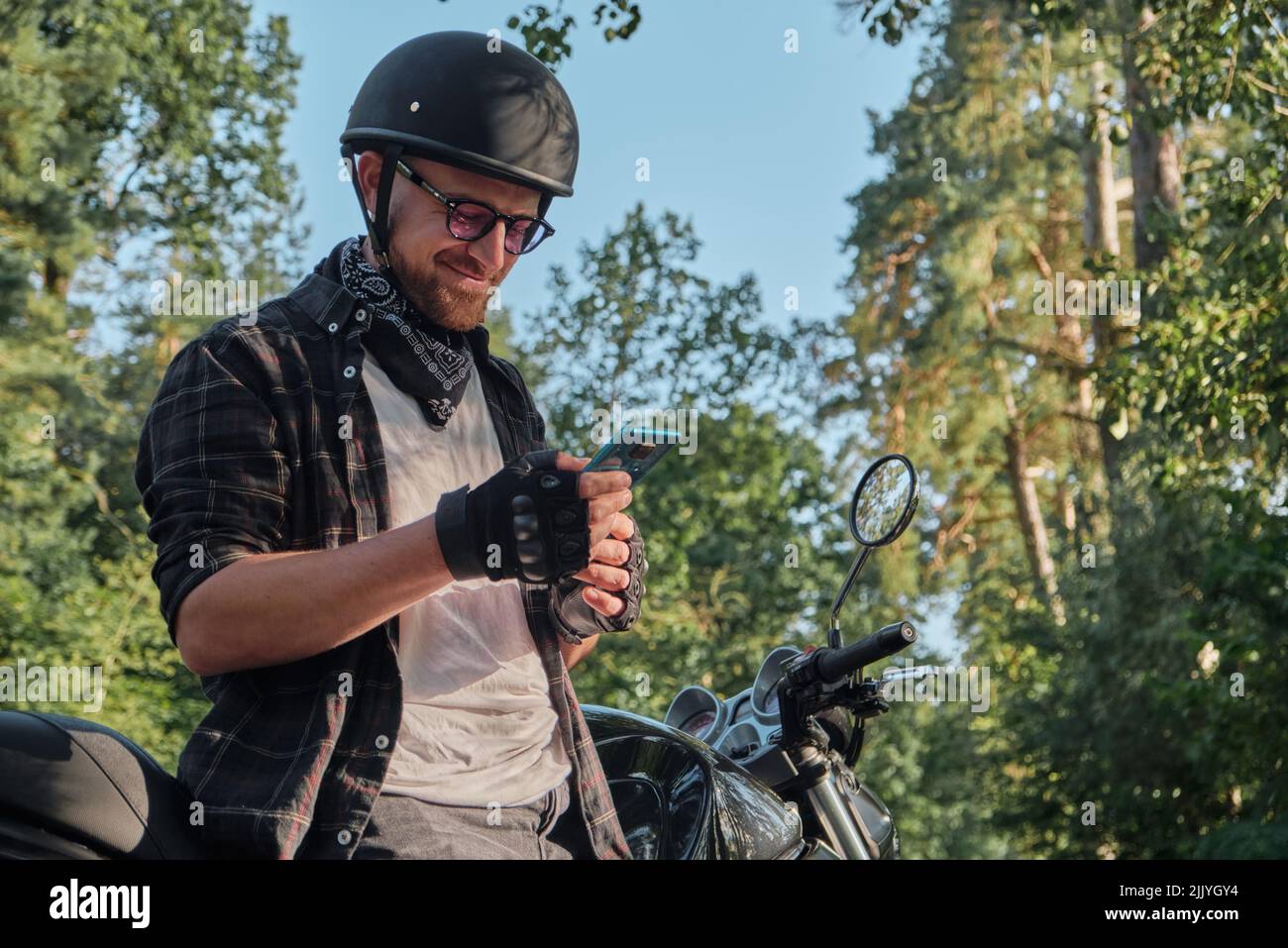 Jeune homme motard dans un casque avec un téléphone portable et souriant assis sur une moto Banque D'Images